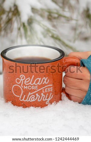 Hand holding hot mug steam rising relax more worry less on front surrounded by snowy scene & icy pine branches in winter background, holiday stress keep calm relax more worry less concept, vertical 