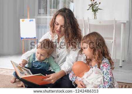 Young mother or nanny with small children, a boy and a girl, sit on the floor on a rug in the room at home and read a book
