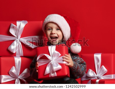 Smiling funny child in Santa red hat holding Christmas gift in hand over red wall background. Christmas eve concept.