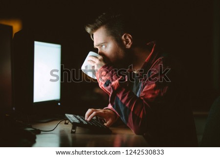 Young handsome man drinking coffee in front of the computer, working late in the dark office