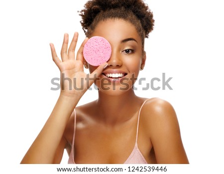 Funny girl holding pink sponge near her face. Portrait of young african american girl on white background. Youth and skin care concept