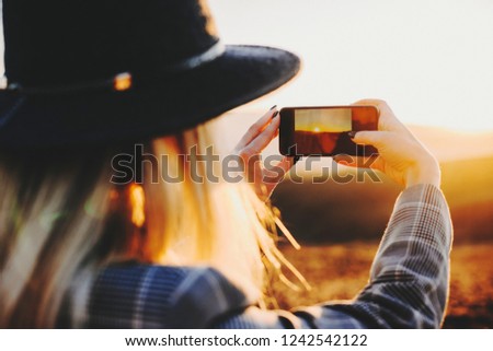Back view of young female using smartphone to take pictures of setting sun in wonderful countryside.Anonymous woman taking pictures of sunset