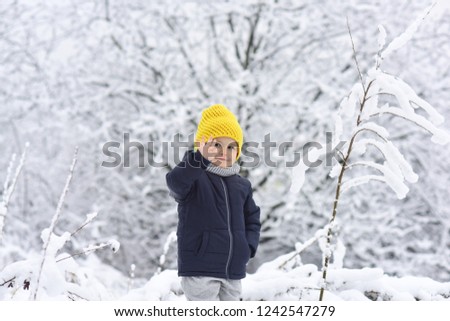 Hello, winter! Handsome boy in black warm jacket and yellow knitted cap. Kid in snow waving hand hello. Friendly boy smiles in the winter forest. Happy childhood. Positive children. Winter walk