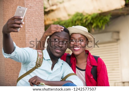 couple of young tourists sitting outdoors photographing themselves with mobile phone while smiling.