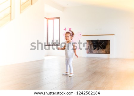 
Beautiful charming little girl wearing fairy costume with magic wand, dancing in a modern house interior, with white background and brown wood floor. Natural light. Horizontal framing.