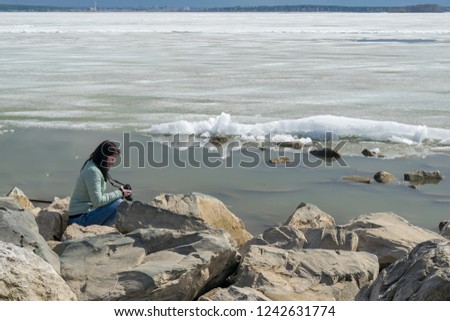 A girl in a warm jacket with a camera on the rocky shore of an ice-covered pond, taking pictures of the landscape