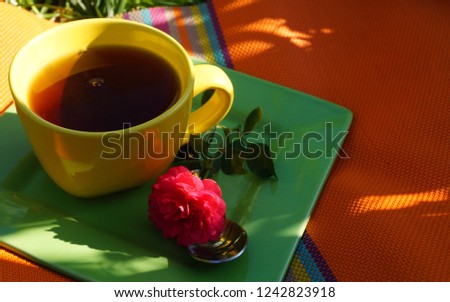 One yellow cup of tea on green plate with orange napkin with a pink rose and an iron spoon in the garden with green grass. Good morning.