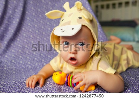 cute baby with blue eyes in yellow bright giraffe costume on blue background in white flower, baby has bright color toy