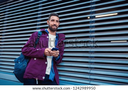 Confident bearded man holding in and mobile phone while standing near street wall in sunny summer evening.