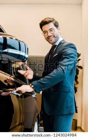 cheerful male car dealer in eyeglasses pointing at black automobile in car salon