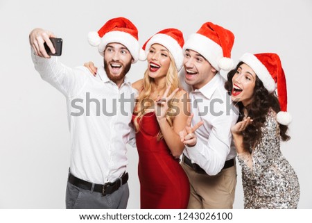 Group of happy friends dressed in red hats standing isolated over white background, celebrating New Year, taking a selfie with mobile phone