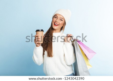 Happy young woman wearing sweater standing isolated over blue background, carrying shopping bags, drinking coffee