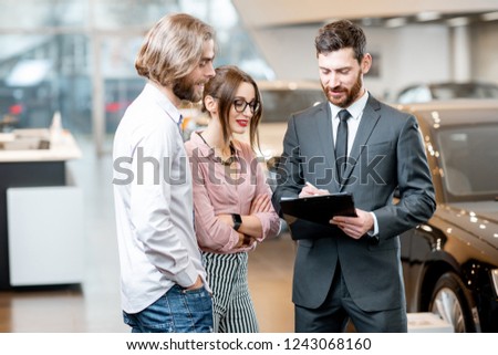 Handsome male dealer selling to a young couple clients a new car, standing together with some documents in the showroom