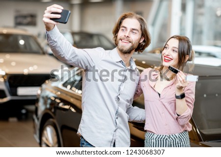 Happy couple making selfie photo holding keys in front of their new car in the showroom