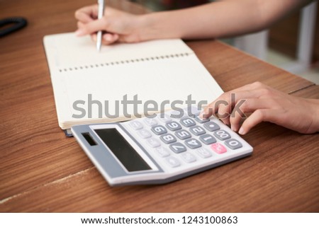 Faceless shot of salon receptionist at wooden desk using calculating machine and writing in notepad
