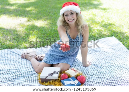Happy woman sitting on grass and holding Christmas gift. Beautiful lady wearing Santa hat and looking at camera. Christmas and nature concept. Front view.