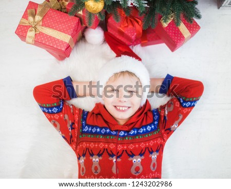 Portrait of little european kid laying on white carpet on floor near christmas tree and holiday presents smiling happily and looking at camera. Hands behind his head in santa red hat.