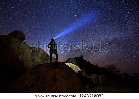 Silhouette of lone woman on rounded rocky structures gazing at milky way and the stars,  focused beam of headlamp pointing directly to milky way. Night sky photo. Twyfelfontein, Damaraland, Namibia. Royalty-Free Stock Photo #1243218085