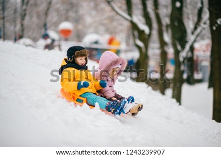 Two small cheerful children ride Board on snow hill on clear frosty day. Brother and sister playing outside in winter. Sincere children's emotions in snow