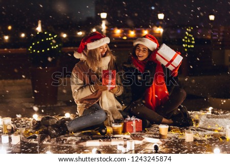 Photo of an excited emotional young friends sitting outdoors in evening in christmas hats holding gift box.