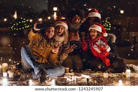 Photo of a happy young group of friends sitting outdoors in evening in christmas hats drinking coffee take a selfie.