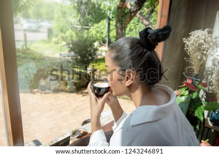 Charming Woman with Beautiful Smile Rest in Coffee Shop, Happy Asian Female Enjoying Morning Sunshine with Black Cup of Cappuccino Coffee while Relaxing by the High Window in Cafe on Free Time