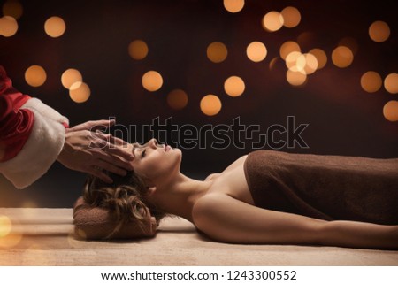 New Year SPA concept. Santa masseur make SPA treatment for young beautiful woman. Warm bokeh lights on background. Royalty-Free Stock Photo #1243300552