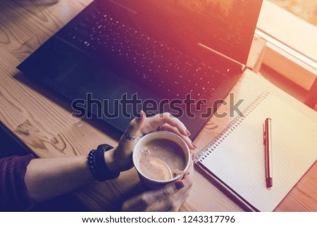 Top view. Student woman holds with two hands a hot cup of tea with a slice of orange. Workplace, notebook with pen and laptop, the working process. coffee shop, warm color toning
