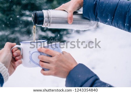 Hands with cups of tea and thermos flask in snowy winter forest close up image