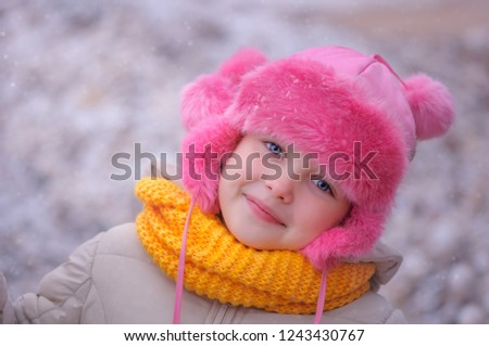 Close View Of Funny Young Girl. Smiling Child In A Fur Pink Hat And A Knitted Yellow Scarf Loose In Winter With snow.