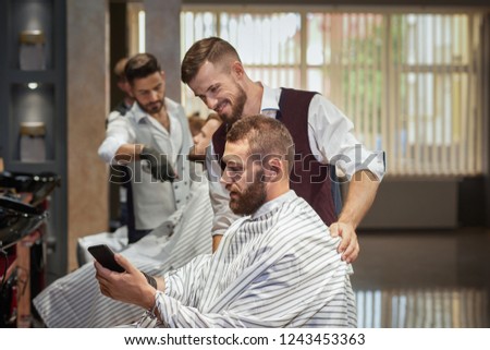 Bearded man sitting in chair in barber shop and showing stylish haircut in phone. Handsome male hairdresser in uniform smiling and looking at picture. Concept of styling of hair and beard.