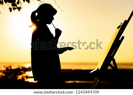 silhouette of a young woman painting a picture with paints on canvas on an easel on nature, girl figure with brush and artist's palette engaged in art at sunset on horizon, concept leisure activity