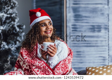 A young beautiful curly girl, wearing a Santa Clause hat and wrapped in a warm blanket, drinks hot coffee on the bed in a loft room. new year \ christmas concept.
