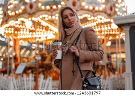 Young pretty woman in a coat and a vintage knitted sweater with black bag holds coffee walks in an amusement park on the background of a carousel decorated with festive garland in the city