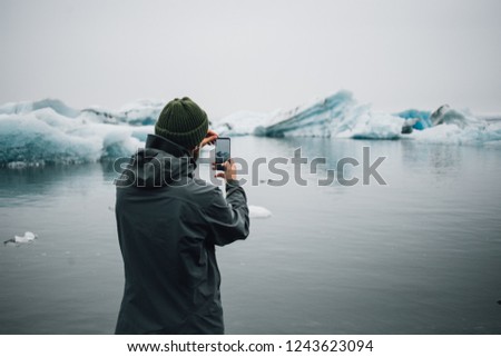 Young hipster or millennial adventurer with futuristic high technology all screen smartphone makes photo or video of impressive melting glaciers or icebergs in ice cold ocean water.