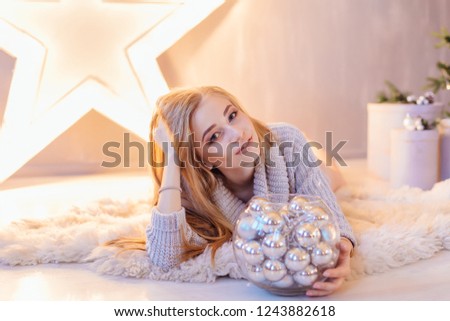 Beautiful young blond woman dressed in white sweater laying next to the big star with bowl full of silver balls in a holiday interior with Christmas tree.