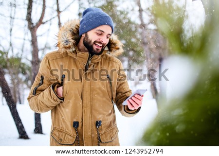 Smiling bearded man wears warm winter clothes and using smartphone with fast internet data connection in country side. Handsome man texting with cellphone and using apps in forest. Snowfall in woods.