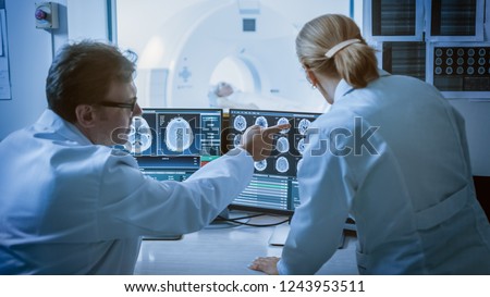 In Control Room Doctor and Radiologist Discuss Diagnosis while Watching Procedure and Monitors Showing Brain Scans Results, In the Background Patient Undergoes MRI or CT Scan Procedure. Royalty-Free Stock Photo #1243953511