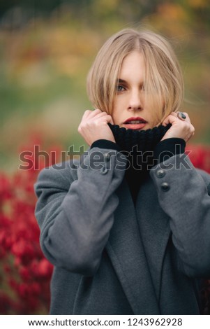 Photo of young woman in gray coat on blurred background of red plants of city