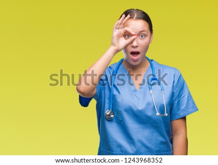 Young caucasian doctor woman wearing medical uniform over isolated background doing ok gesture shocked with surprised face, eye looking through fingers. Unbelieving expression.