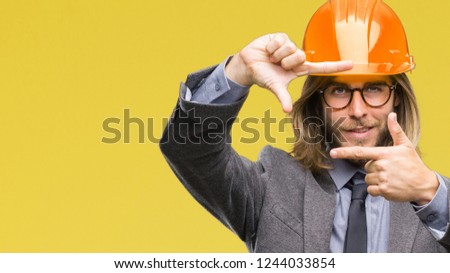 Young handsome architec man with long hair wearing safety helmet over isolated background smiling making frame with hands and fingers with happy face. Creativity and photography concept.