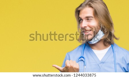 Young handsome doctor man with long hair over isolated background smiling with happy face looking and pointing to the side with thumb up.