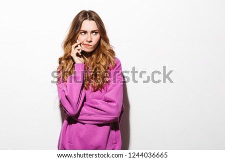 Portrait of upset woman 20s wearing sweatshirt talking on smartphone while standing isolated over white background