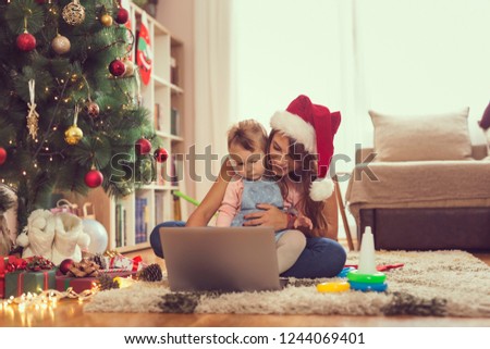 Mother and daughter sitting on the floor next to a Christmas tree, watching cartoons on a laptop computer and having fun. Focus on the baby girl
