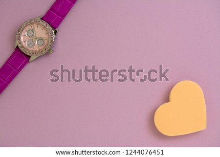 Beautiful women's wrist watch and heart shaped makeup sponge on purple background.