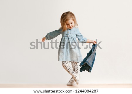 Full length portrait of cute little kid girl in stylish jeans clothes looking at camera and smiling, standing against white studio wall. Kids fashion concept Royalty-Free Stock Photo #1244084287