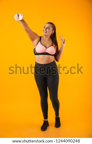 Full length portrait of caucasian chubby woman in sportive bra taking selfie photo on mobile phone isolated over yellow background