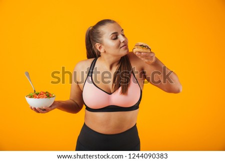 Portrait of a cheerful overweight fitness woman wearing sports clothing standing isolated over yellow background, holding bowl with salad and a burger