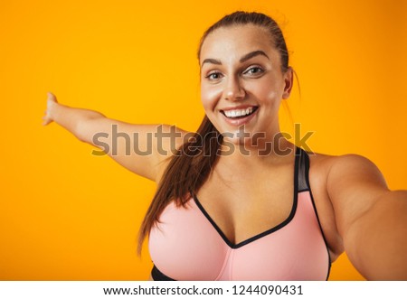 Portrait of a cheerful overweight fitness woman wearing sports clothing standing isolated over yellow background, taking a selfie with mobile phone