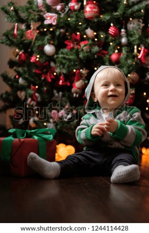 Happy baby in red pajamas on Christmas morning unpacks presents in front of the Christmas tree on a dark wooden floor
